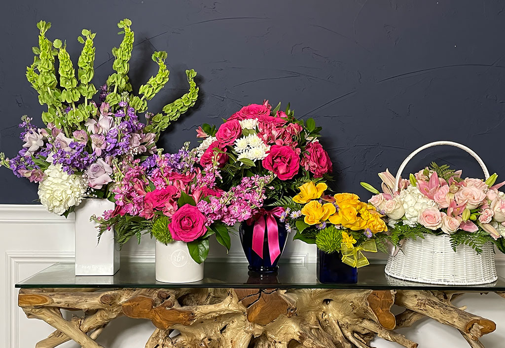 Colorful flower arrangements on a glass table with driftwood base, featuring pink roses, yellow spray roses, purple stock, bells of Ireland, and soft pastel blooms, designed by Ritzy Blooms in Fort Lauderdale.