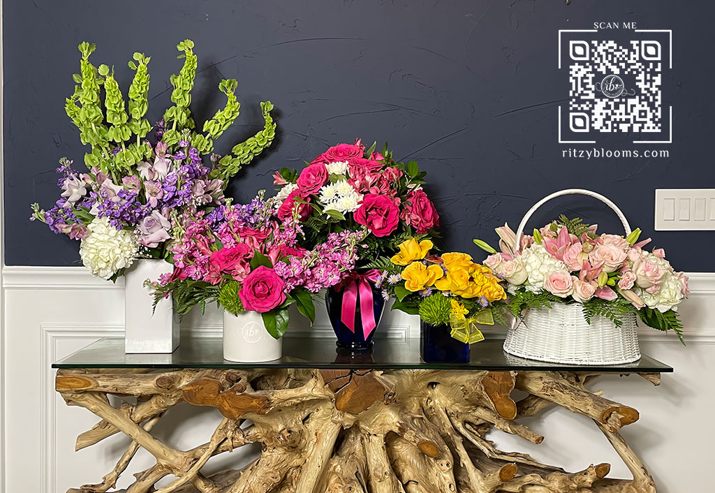 A collection of fresh floral arrangements displayed on a rustic driftwood console table against a navy blue wall. The flowers include hot pink roses, green bells of Ireland, white hydrangeas, yellow calla lilies, soft pink roses, and lilac blooms. A QR code linking to ritzyblooms.com is visible in the top right corner.