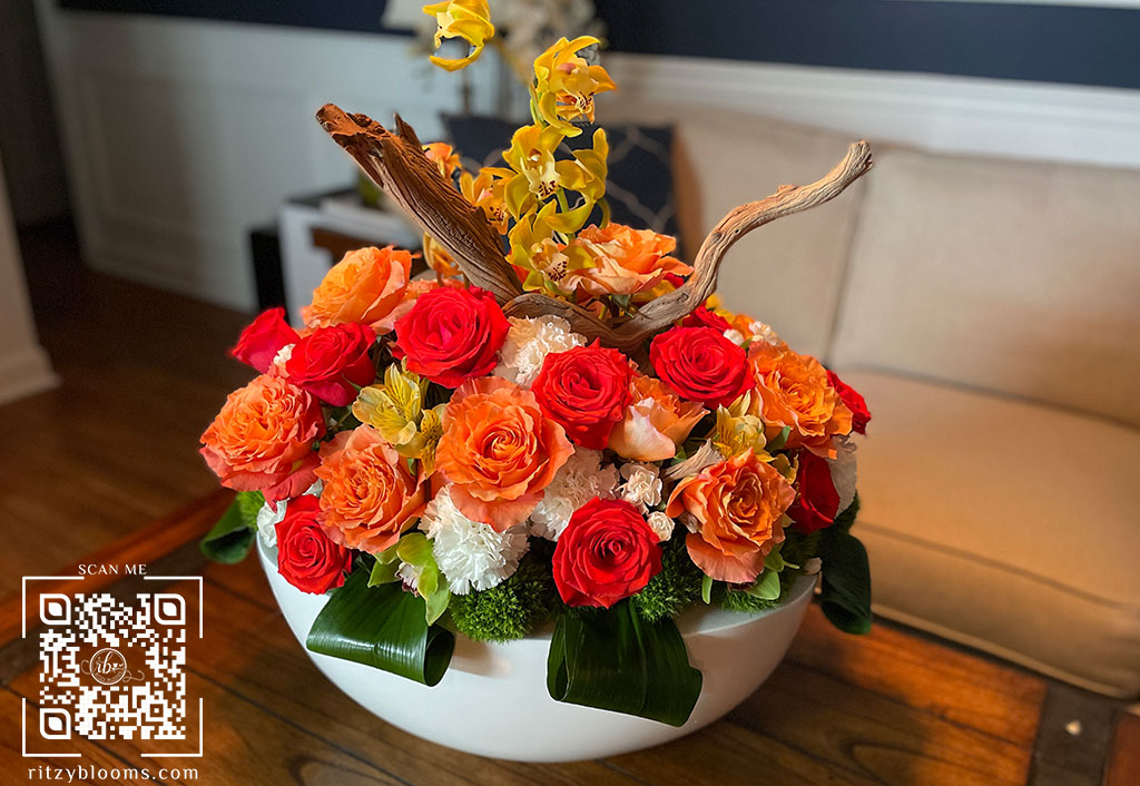 Elegant hotel-style floral arrangement featuring orange, red, and peach roses with white hydrangeas, yellow orchids, and natural driftwood accents, displayed in a modern white bowl by Ritzy Blooms in Fort Lauderdale.